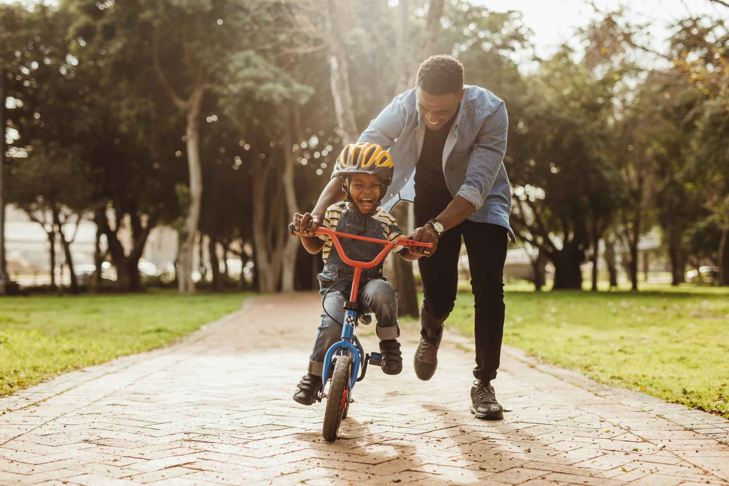 A father helps his laughing child ride a bicycle along a tree-lined path.