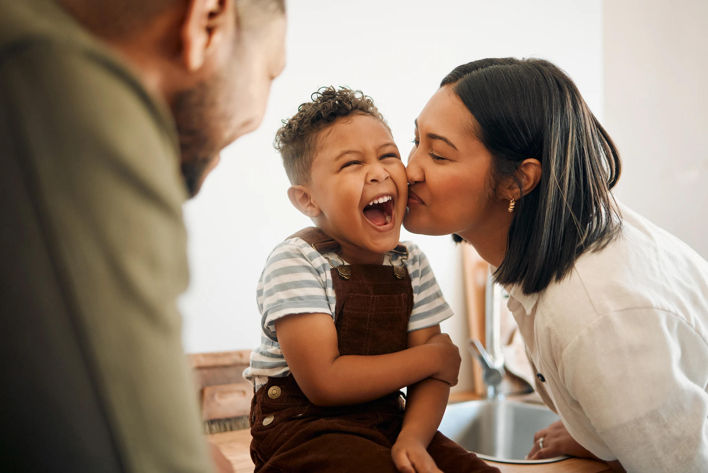 A child laughs as their mother kisses their cheek, with their father close beside them.
